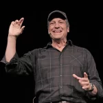 Event photo of Mike Rowe on stage with his right hand raised as he tells story of being in the sewer.