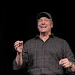 Mike Rowe wearing a baseball cap on stage during a keynote speech to a group of service workers